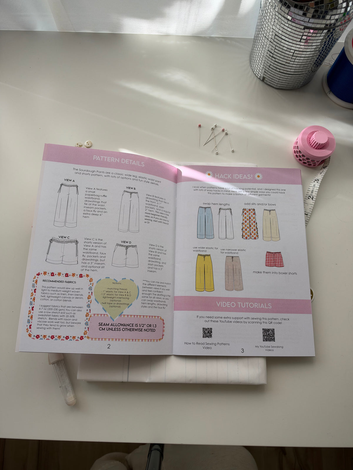 An open sewing pattern booklet showing pattern details and sketch ideas for shorts, placed on a white desk with sewing supplies nearby.
