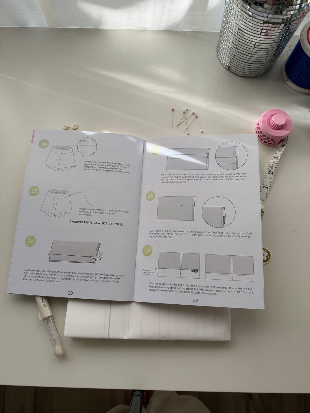 An open instruction booklet showing step-by-step sewing or crafting diagrams laid on a white desk, surrounded by sewing supplies including pins, a pink spool, and a silver cup.