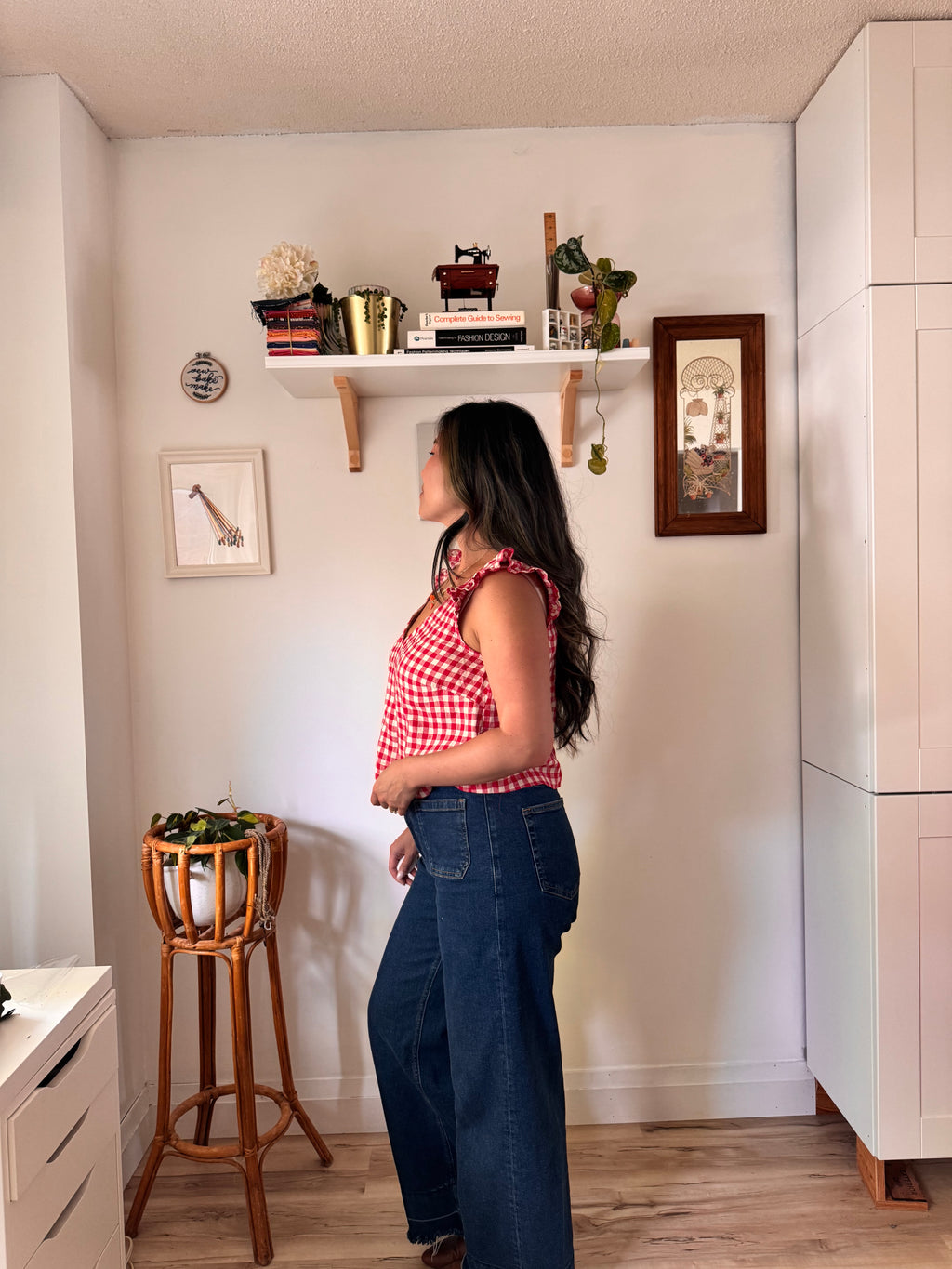 A woman wearing a red and white gingham ruffled sleeveless top and dark blue wide-leg jeans stands in profile in a room, looking up at a white floating shelf with books and decor.